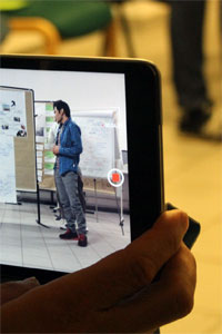 Foto eines Tablets, auf welchem die Kamerafunktion geöffnet ist. Das Display zeigt einen Erwachsenen vor einer Pinnwand.