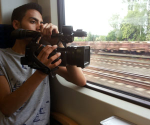 Ein junger Mann sitzt in einem Zug und filmt mit einer Videokamera aus dem Fenster.