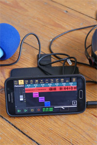 Auf dem Display eines Smartphones sind mehrere Tonspuren abgebildet. Neben dem Smartphone liegen ein Mikrofon und Kopfhörer.