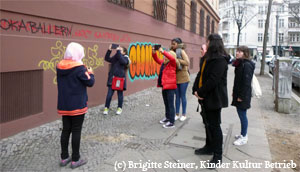 Mehrere Personen stehen vor einer Hauswand, auf welche verschiedene Graffitis gesprüht wurden und betrachten diese. Ein paar der Personen fotografieren die Wand mit ihren Smartphones.