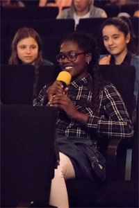 Bild eines Mädchens, das in einem Kinosessel sitzt. Es lächelt und hält ein Mikrofon in den Händen.