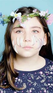 Ein Mädchen trägt einen Blumenkranz auf dem Kopf und schaut direkt in die Kamera. Auf seinen Wangen liegen Zwiebelspalten.