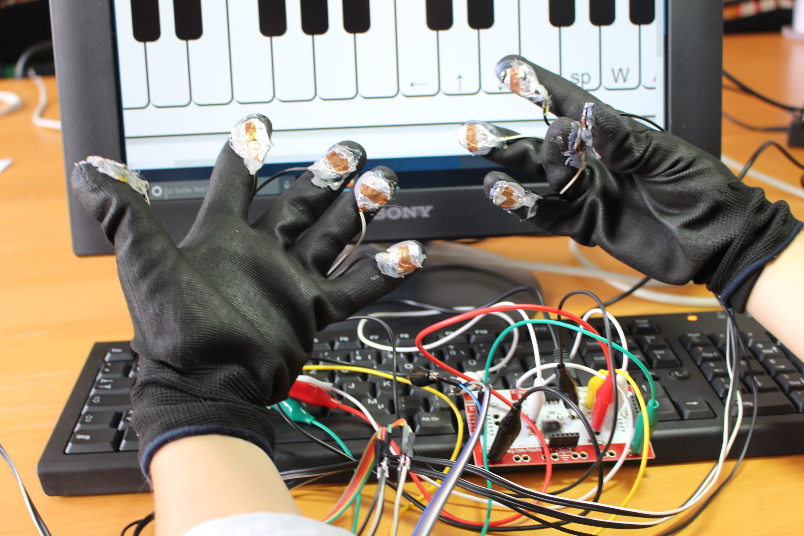 Zwei Hände mit Klavierhandschuhen werden vor einen Laptop mit einer Klavier-App darauf gehalten.