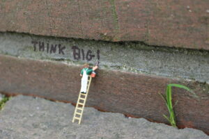 Ein Miniaturmännchen auf einer Leiter schreibt "Think big!" auf eine Wand.