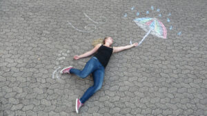 Ein Mädchen liegt auf dem Boden. Über ihr ist ein Schirm mit Kreide auf den Boden gemalt. das Foto ist von oben aufgenommen worden, sie scheint mit dem Schirm in der Hand zu fliegen.
