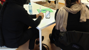 Zwei Menschen von hinten an einem Tisch sitzend; in der Mitte ein Smartphone